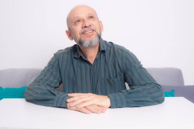 Kel, sakallı, yeşil gömlekli, kolları masada dinlenen, gülümseyen, sakin ve rahat bir adam. Beyaz arkaplana karşı.