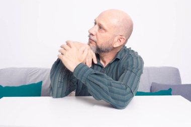 Ciddi, resmi tişörtlü, sakallı, kollarını masaya uzatmış, düşünceli ve sakin bir adam. Beyaz arkaplana karşı.