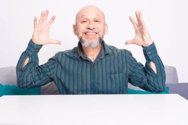 Yeşil tişörtlü kel, sakallı bir adam, kolları masanın üzerinde dinleniyor, el kol hareketi yapıyor ve sakince gülümsüyor ve rahatlıyor. Beyaz arkaplana karşı.