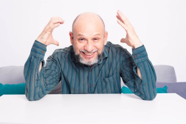 Yeşil tişörtlü kel, sakallı bir adam, kolları masanın üzerinde dinleniyor, el kol hareketi yapıyor ve sakince gülümsüyor ve rahatlıyor. Beyaz arkaplana karşı.