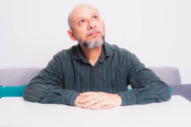 Ciddi, resmi tişörtlü, sakallı, kollarını masaya uzatmış, düşünceli ve sakin bir adam. Beyaz arkaplana karşı.