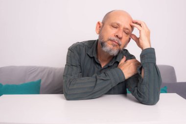 Kel, iyi giyimli, sakallı bir adam, kollarını masaya uzatmış, düşünceli ve sakin bir şekilde oturuyor. Beyaz arkaplana karşı.