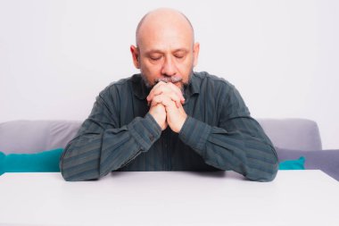 Ciddi, resmi tişörtlü, sakallı, kollarını masaya uzatmış, düşünceli ve sakin bir adam. Beyaz arkaplana karşı.