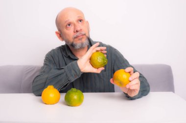 Elinde iki mandalina tutan yeşil gömlekli, sakallı kel bir adam. Beyaz arkaplana karşı.