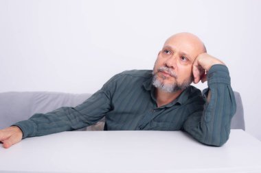 Kel, iyi giyimli, sakallı bir adam, kollarını masaya uzatmış, düşünceli ve sakin bir şekilde oturuyor. Beyaz arkaplana karşı.