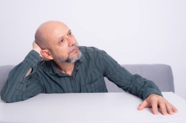 Kel, iyi giyimli, sakallı bir adam, kollarını masaya uzatmış, düşünceli ve sakin bir şekilde oturuyor. Beyaz arkaplana karşı.
