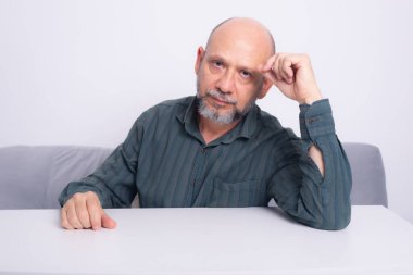 Ciddi, resmi tişörtlü, sakallı, kollarını masaya uzatmış, düşünceli ve sakin bir adam. Beyaz arkaplana karşı.