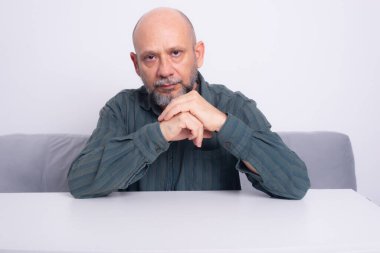 Ciddi, resmi tişörtlü, sakallı, kollarını masaya uzatmış, düşünceli ve sakin bir adam. Beyaz arkaplana karşı.