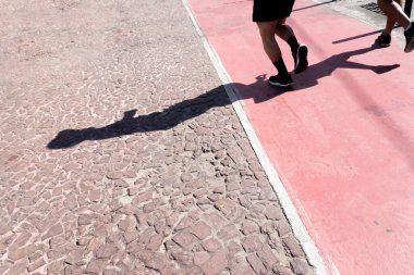 Bir insanın alt kısmı ve gölgesi, güçlü güneş ışığı altında sahil boyunca koşuyor. Sağlıklı spor pratiği. Brezilya.