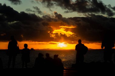 Siluetli figürler, güneş ufuktan batarken ahşap bir iskelede toplanır ve suyun üzerinde sıcak bir parıltı oluştururlar..