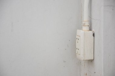 Beyaz bir duvara elektrik prizi monte edilmiş. Prizin iki yuvası ve duvarın tepesine çıkan bir güç kablosu var. Duvarda çatlaklar ve lekeler var..