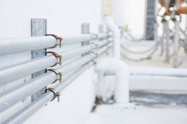 Beyaz bir duvara birkaç metal boru monte edilmiştir. Bu borular metal kıskaçlarla tutturulmuş. Duvara tutturulmuş metal bir levhaya tutturulmuş..