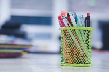 Kalemler, kalemler ve fosforlu kalemler gibi çeşitli yazma araçları içeren yeşil ağ kalem tutucu. Arkaplan, bilgisayar monitörü de dahil olmak üzere bulanık ögelerle dolu bir ofis ayarına benziyor..