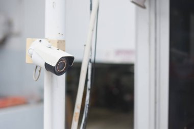 Güvenlik kamerası beyaz bir direğe monte edilmiş. Kamera çevreyi gözetlemek için yerleştirilmiş. Binalar ve arka planda çitlerle çevrili gibi görünüyor.