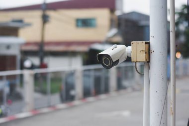 Güvenlik kamerası beyaz bir direğe monte edilmiş. Kamera çevreyi gözetlemek için yerleştirilmiş. Binalar ve arka planda çitlerle çevrili gibi görünüyor.