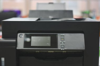 Ofis ortamında bir masanın üzerine yerleştirilmiş siyah çoklu işlevli bir yazıcı. Bu resim günlük görev tanımlama, tarama ve belgelerin kopyalanması için gerekli olan ortak ofis ekipmanlarını tasvir ettiği için alakalıdır..