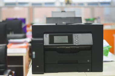 Ofis ortamında bir masanın üzerine yerleştirilmiş siyah çoklu işlevli bir yazıcı. Bu resim günlük görev tanımlama, tarama ve belgelerin kopyalanması için gerekli olan ortak ofis ekipmanlarını tasvir ettiği için alakalıdır..