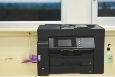 Ofis ortamında bir masanın üzerine yerleştirilmiş siyah çoklu işlevli bir yazıcı. Bu resim günlük görev tanımlama, tarama ve belgelerin kopyalanması için gerekli olan ortak ofis ekipmanlarını tasvir ettiği için alakalıdır..