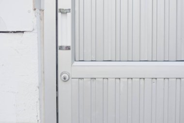 Dikey çıkıntıları olan metalik bir kapının yakın görüntüsü. Kapının sol tarafında anahtar deliği ve sapı var. Kapı beyaz dış duvarı olan bir binanın parçası gibi görünüyor..