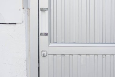 Dikey çıkıntıları olan metalik bir kapının yakın görüntüsü. Kapının sol tarafında anahtar deliği ve sapı var. Kapı beyaz dış duvarı olan bir binanın parçası gibi görünüyor..