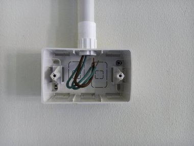 Duvara monte edilmiş elektrik bağlantı kutusunu aç. Kutu beyaz ve dikdörtgen, üstten giren bir kanal var. Kutunun içinde üç kablo var: iki yeşil kablo ve bir kahverengi kablo..