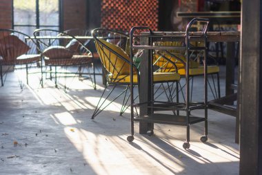 İçinde minderleri olan metal çerçeveli sandalyeler olan kapalı bir oturma odası. Sandalyeler masaların etrafına dizilir, ve güneş ışığı büyük pencerelerden içeri akar, yerde gölgeler bırakır..