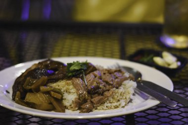 Metal bir ağ masasında bir tabak yemek. Tabakta, haşlanmış et ve sebze gibi görünen bazı otlarla süslenmiş pirinçler var..