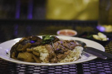 Metal bir ağ masasında bir tabak yemek. Tabakta, haşlanmış et ve sebze gibi görünen bazı otlarla süslenmiş pirinçler var..