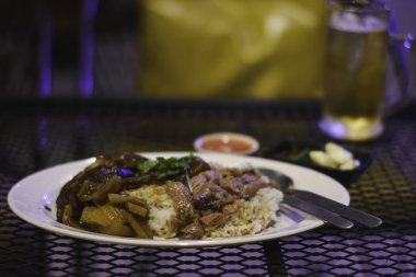 Metal bir ağ masasında bir tabak yemek. Tabakta, haşlanmış et ve sebze gibi görünen bazı otlarla süslenmiş pirinçler var..