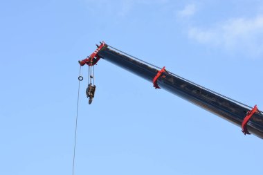 Bir vincin üst kısmı açık mavi gökyüzüne karşı. Turnanın kolu çapraz olarak uzatılmış ve bir kanca vincin kablosunun ucunda asılı..