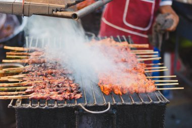 Bir sokak yemeği satıcısı ızgarada tavuk şişi pişiriyor. Şişler sıra sıra dizilir, bazıları koyu, kömürleşmiş etle, diğerleri hafif, turuncu etle..