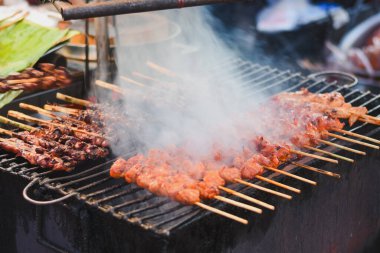 Bir sokak yemeği satıcısı ızgarada tavuk şişi pişiriyor. Şişler sıra sıra dizilir, bazıları koyu, kömürleşmiş etle, diğerleri hafif, turuncu etle..