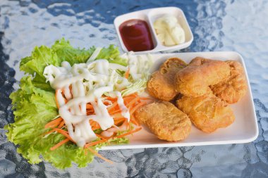 Tabakta birkaç altın-kahverengi kızarmış tavuk nugget, yanında maruldan oluşan taze salata ve beyaz bir sos serpiştirilmiş lahana bulunuyor.. 