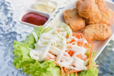 Tabakta birkaç altın-kahverengi kızarmış tavuk nugget, yanında maruldan oluşan taze salata ve beyaz bir sos serpiştirilmiş lahana bulunuyor.. 