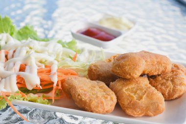 Tabakta birkaç altın-kahverengi kızarmış tavuk nugget, yanında maruldan oluşan taze salata ve beyaz bir sos serpiştirilmiş lahana bulunuyor.. 