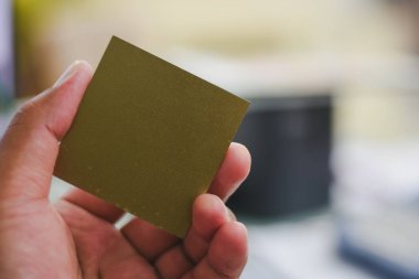 Küçük, kare, metalik görünümlü bir nesneyi tutan bir el. Nesne ince, düz bir madde gibi görünüyor, pürüzsüz, yansıtıcı bir yüzeyi var, muhtemelen metal ya da metalik kaplamalı bir madde..