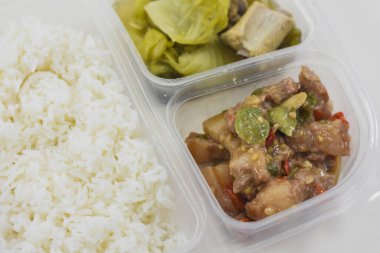 Plastik bir kabın içinde üç bölmeli bir yemek. Üst bölümde beyaz pirinçler var. Sağ alt bölümde bir çorba ya da kemik içi et ve yapraklı yeşil bir güveç var..