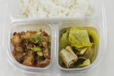 Plastik bir kabın içinde üç bölmeli bir yemek. Üst bölümde beyaz pirinçler var. Sağ alt bölümde bir çorba ya da kemik içi et ve yapraklı yeşil bir güveç var..