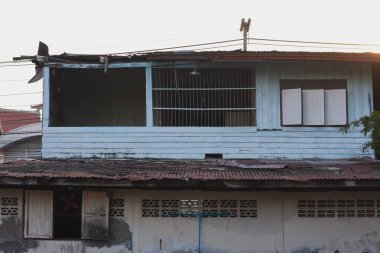 Ahşap kaplamalı iki katlı bir bina. Üst katta açık mavi ahşap duvarlar var. Ortasında dikey metal parmaklıklar var. Muhtemelen bir pencere ya da balkon alanı var..