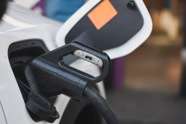 Şarj kapağı açık beyaz bir elektrikli aracın yan tarafında ve şarj kablosu takılı. Şarj portu aracın ön tarafında, yan aynanın hemen arkasında..