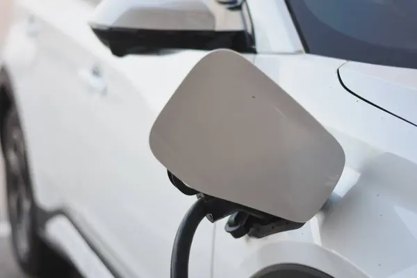 Şarj kapağı açık beyaz bir elektrikli aracın yan tarafında ve şarj kablosu takılı. Şarj portu aracın ön tarafında, yan aynanın hemen arkasında..