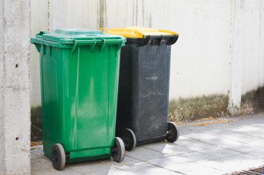 Duvarın yanındaki kaldırımda duran iki büyük tekerlekli çöp tenekesi. Bir kutu yeşil ve yeşil kapaklı, diğeri ise sarı kapaklı siyah..