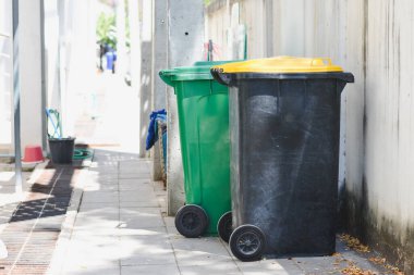 Duvarın yanındaki kaldırımda duran iki büyük tekerlekli çöp tenekesi. Bir kutu yeşil ve yeşil kapaklı, diğeri ise sarı kapaklı siyah..