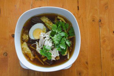 Bir kase çorba ahşap bir masada oturur. Çorba, kaynamış yumurta, doğranmış tavuk, taze kişniş yaprakları ve bambu filizlerine ya da diğer sebzelere benzeyen malzemeler içerir..