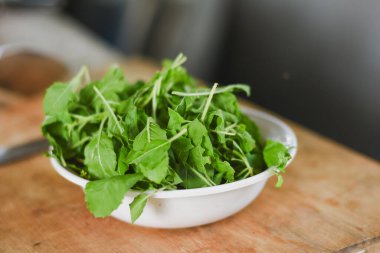 Beyaz bir kâseye odaklanan tepeden çekimler. Açık ahşap bir yüzeye yerleştirilmiş taze yeşil yapraklarla dolu..
