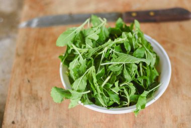 Beyaz bir kâseye odaklanan tepeden çekimler. Açık ahşap bir yüzeye yerleştirilmiş taze yeşil yapraklarla dolu..