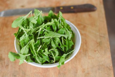 Beyaz bir kâseye odaklanan tepeden çekimler. Açık ahşap bir yüzeye yerleştirilmiş taze yeşil yapraklarla dolu..