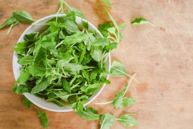 Beyaz bir kâseye odaklanan tepeden çekimler. Açık ahşap bir yüzeye yerleştirilmiş taze yeşil yapraklarla dolu..