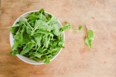 Beyaz bir kâseye odaklanan tepeden çekimler. Açık ahşap bir yüzeye yerleştirilmiş taze yeşil yapraklarla dolu..