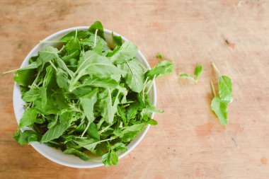 Beyaz bir kâseye odaklanan tepeden çekimler. Açık ahşap bir yüzeye yerleştirilmiş taze yeşil yapraklarla dolu..